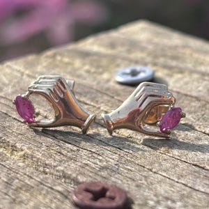 May include: A pair of silver-toned earrings, each shaped like a hand holding a pink gemstone. The earrings are set against a weathered wooden surface, with a blurred background.