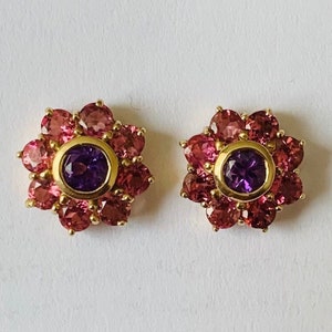 K&ouml;nnte beinhalten: Ein Paar goldene Ohrringe mit einem violetten Edelstein, der von rosa Edelsteinen in Bl&uuml;tenform umgeben ist.