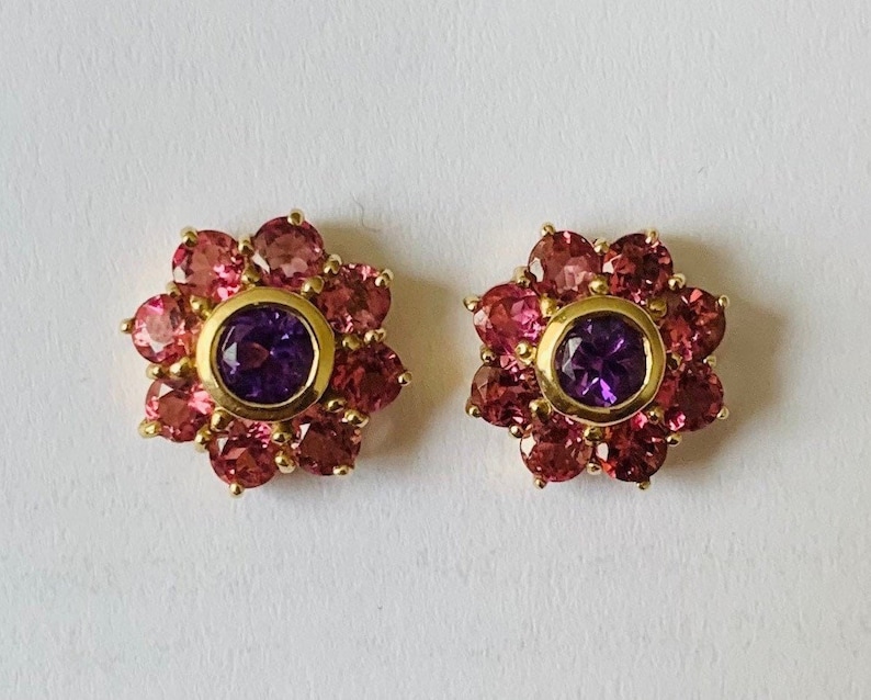 K&ouml;nnte beinhalten: Ein Paar goldene Ohrringe mit einem violetten Edelstein, der von rosa Edelsteinen in Bl&uuml;tenform umgeben ist.