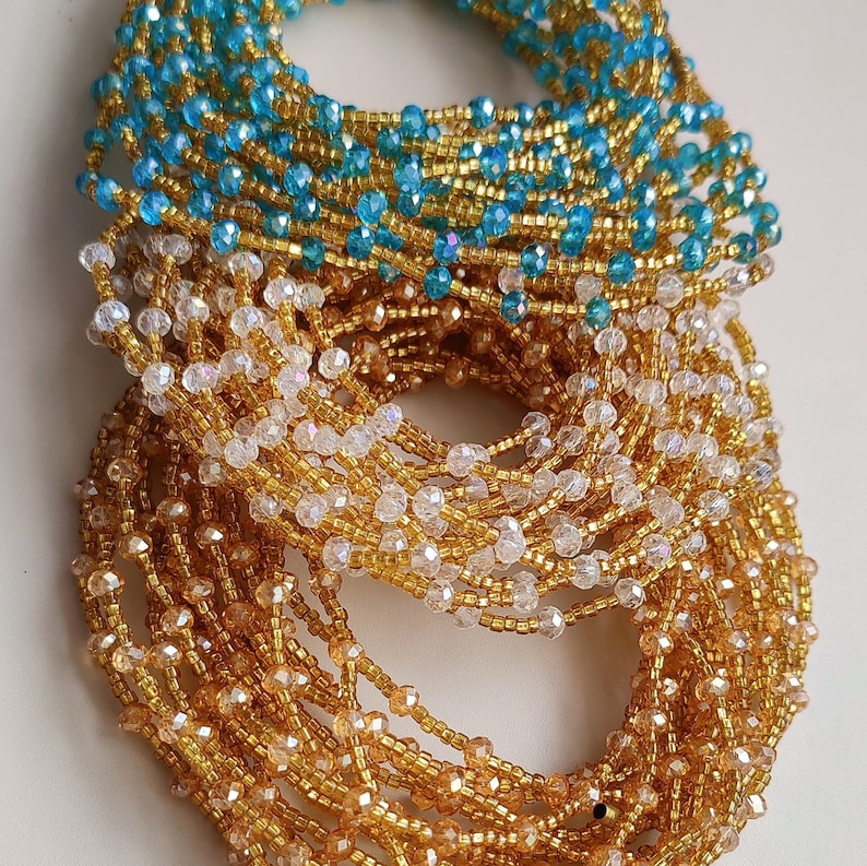 May include: A stack of beaded waist beads in gold, clear, and blue colors. The beads are strung together in a continuous strand and are arranged in a circular pattern.