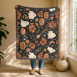 May include: Black throw blanket with a Halloween-themed pattern. The design includes white ghosts, orange pumpkins, flowers, and crescent moons. The blanket has a fringed edge and is held up by a person.