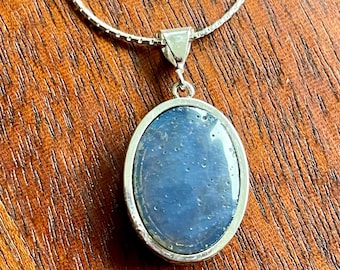 Può includere: Una collana color argento con un ciondolo in pietra blu di forma ovale e superficie liscia, incastonato in una cornice di metallo lucido. Il ciondolo pende da una sottile catena testurizzata che cattura la luce, creando un riflesso delicato.