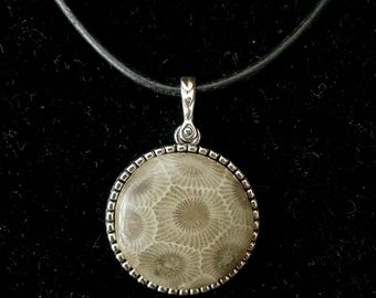 Può includere: Un ciondolo rotondo con una pietra di corallo fossilizzato dal motivo floreale beige e grigio, incastonato in una montatura in metallo argentato testurizzato. Il ciondolo pende da un cordino nero liscio, creando un punto focale circolare.