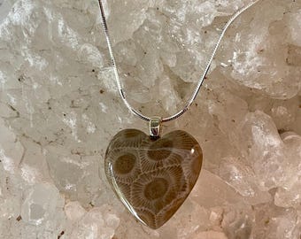 Può includere: Un ciondolo a forma di cuore realizzato in corallo fossile marrone lucido con un distinto motivo circolare, sospeso a una sottile catena a serpente color argento. Il ciondolo presenta una finitura liscia e lucida ed è fissato a una semplice contromaglia.