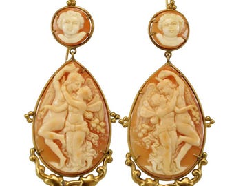 May include: A pair of gold tone earrings with cameo carvings of a couple embracing. The earrings feature a teardrop-shaped cameo with a detailed carving of a couple embracing, surrounded by a gold tone frame. The earrings also feature a smaller round cameo with a carving of a single face, also surrounded by a gold tone frame.