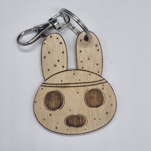 May include: A wooden keychain featuring a cartoon animal with large ears and a round face. The keychain is brown and has a light brown colour. The animal has two large eyes and a small mouth. The keychain is attached to a silver metal ring.