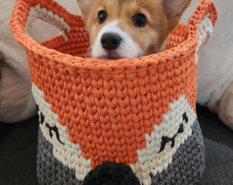 May include: A small, brown and white corgi puppy sits inside a crocheted orange, white, and gray basket with a fox face design.