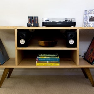 May include: A light wood record player stand with two shelves, a record player, two speakers, and a bowl on the top shelf. There are books on the bottom shelf and vinyl records on either side of the stand.
