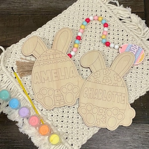 May include: Two unfinished wooden bunny-shaped Easter egg ornaments with the names Amelia and Charlotte written on them. The ornaments are surrounded by colorful paint and a string of beads.