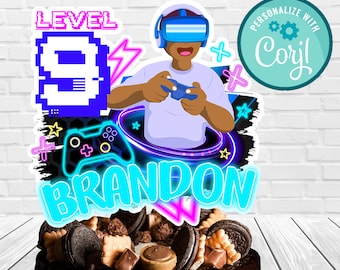 May include: A cake topper featuring a cartoon character wearing a virtual reality headset and holding a video game controller. The topper is decorated with neon colors and the text "LEVEL 9 BRANDON" and "PERSONALIZE WITH Coryl".
