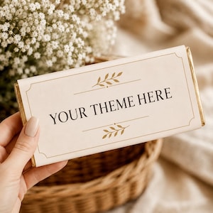 May include: A rectangular chocolate bar wrapper in cream with gold foil edges. The center features the black text "YOUR THEME HERE" framed by thin gold lines and decorative gold leaf motifs above and below the text, creating a clean design.