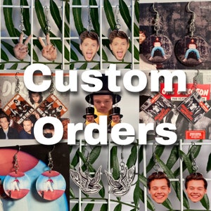 Könnte beinhalten: Individuelle Bestellungen für Ohrringe mit verschiedenen Designs, darunter ein Peace-Zeichen, ein Porträt eines Mannes, ein Vogel und ein Albumcover einer Band. Die Ohrringe werden auf einem Drahtgestell mit grünen Pflanzen im Hintergrund präsentiert.