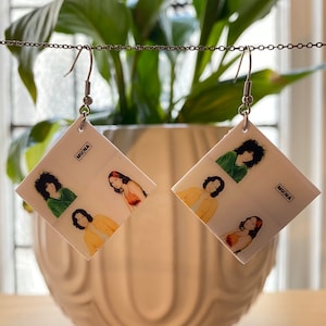 May include: A pair of white square earrings with a band photo of the band MUNA. The earrings are hanging from silver hooks.