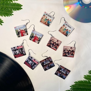 Może przedstawiać: Kolczyki z okładkami albumów zespołu One Direction. Kolczyki przedstawiają okładki albumów z "Midnight Memories" i "Made in the A.M." oraz inne okładki. Kolczyki są kwadratowe ze srebrnymi haczykami. Płyta CD i płyta winylowa znajdują się w tle.