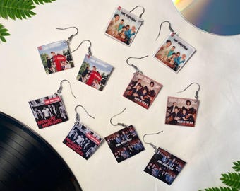 Può includere: Orecchini con copertine di album della band One Direction. Gli orecchini mostrano copertine di album di "Midnight Memories" e "Made in the A.M." e altre copertine. Gli orecchini sono quadrati con ganci argentati. Un CD e un disco in vinile sono sullo sfondo.