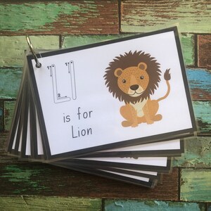May include: A flashcard with a black border and a white background. The letter "L" is written in black with arrows showing the stroke order. A cartoon lion is illustrated on the right side of the card. The text "is for Lion" is written below the letter.