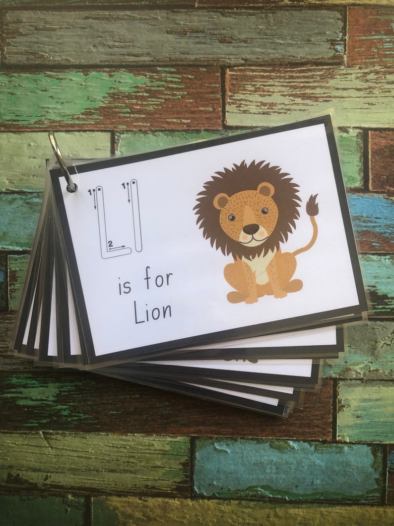 May include: A flashcard with a black border and a white background. The letter "L" is written in black with arrows showing the stroke order. A cartoon lion is illustrated on the right side of the card. The text "is for Lion" is written below the letter.