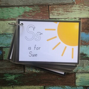 May include: A black and white flashcard with the letter "S" written in cursive and a yellow sun with rays. The text "S is for Sun" is written below the sun.