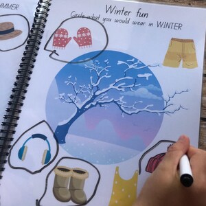 May include: A page from a children's book about seasons. The page is titled "Winter Fun" and asks the reader to circle what they would wear in winter. The page features illustrations of a winter scene, mittens, boots, a hat, and a pair of headphones.