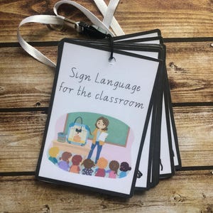May include: A set of flashcards with black borders and a white background. The cards feature illustrations of a teacher and students in a classroom setting. The text on the front card reads "Sign Language for the classroom".