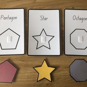 May include: Three white cards with black outlines and black text. The cards show a pentagon, a star, and an octagon. Each shape has a white square in the center. Below the cards are three felt shapes: a red pentagon, a yellow star, and a gray octagon.