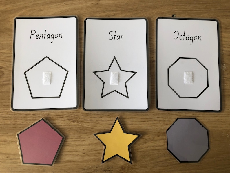 May include: Three white cards with black outlines and black text. The cards show a pentagon, a star, and an octagon. Each shape has a white square in the center. Below the cards are three felt shapes: a red pentagon, a yellow star, and a gray octagon.