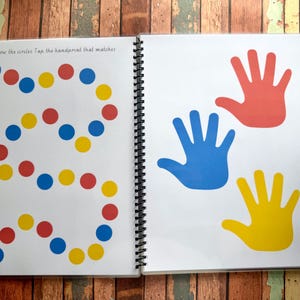 May include: Open spiral-bound activity book with white pages. One page features a winding path of red, blue, and yellow circles. The facing page displays red, blue, and yellow handprints. Text reads: "Follow the circles. Tap the handprint that matches."