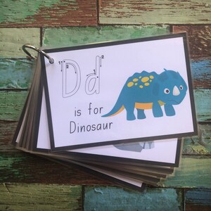 May include: A printable flashcard featuring a blue and yellow dinosaur illustration. The card shows the letter "D" with dotted lines to help trace the letter. The text "D is for Dinosaur" is printed below the illustration.