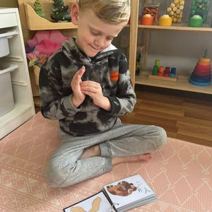 May include: A child sits on a pink and white patterned play mat, holding a book with a picture of a brown and white animal. The book is open to a page with the text "The Y." The child is wearing a black and grey hooded sweatshirt and grey trousers.