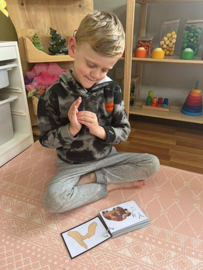 May include: A child sits on a pink and white patterned play mat, holding a book with a picture of a brown and white animal. The book is open to a page with the text "The Y." The child is wearing a black and grey hooded sweatshirt and grey trousers.