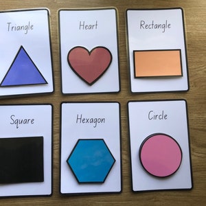 May include: Six white cards with black borders, each featuring a different shape: triangle, heart, rectangle, square, hexagon, and circle. The shapes are filled with different colors: blue, red, orange, black, blue, and pink.