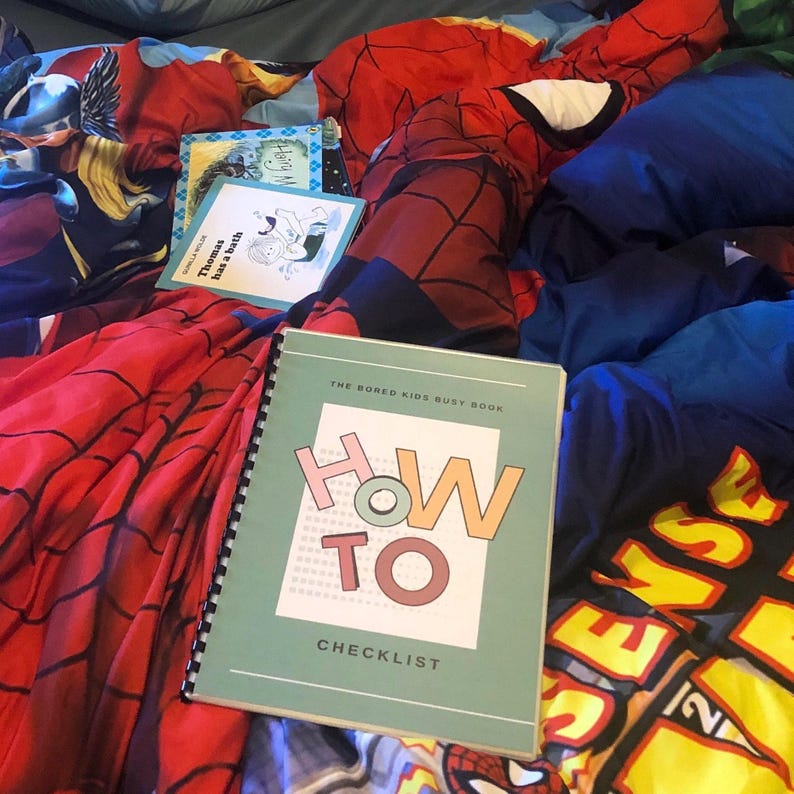 May include: A teal blue book with the title "The Bored Kids Busy Book: How To Checklist" in white and pink lettering. The book is lying on a red and black patterned blanket.
