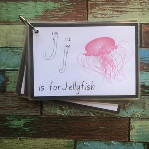 May include: A black and white flashcard with a tracing guide for the letter J. The letter J is traced in black ink. The card also features a pink jellyfish with long tentacles. The text "J is for Jellyfish" is printed below the image.