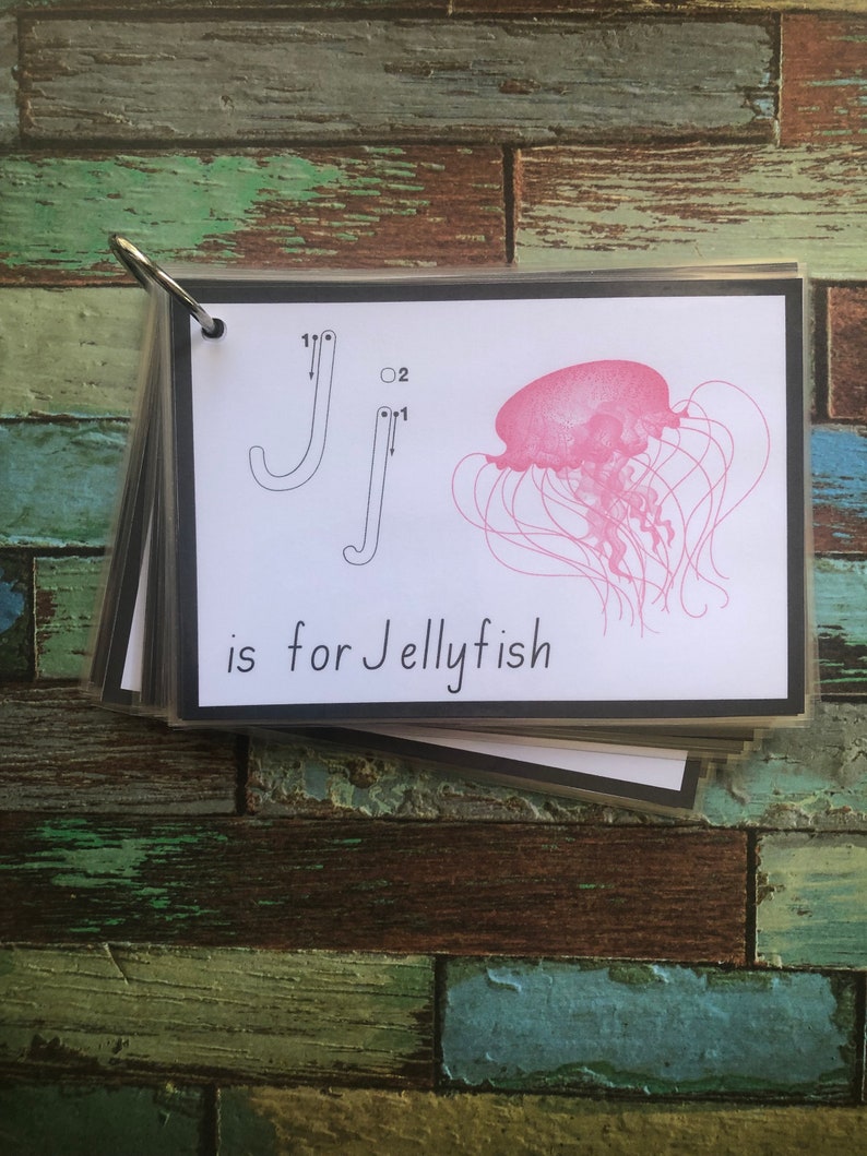 May include: A black and white flashcard with a tracing guide for the letter J. The letter J is traced in black ink. The card also features a pink jellyfish with long tentacles. The text "J is for Jellyfish" is printed below the image.