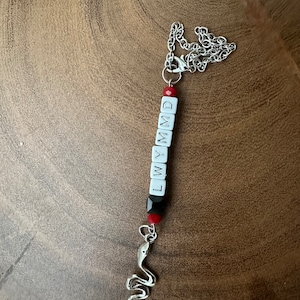 Puede incluir: Un collar de cadena plateada con una cuenta roja y un dije que dice "LWYMMD". El dije está hecho de cuentas blancas con detalles negros. Un dije de serpiente plateada cuelga de la parte inferior del dije.