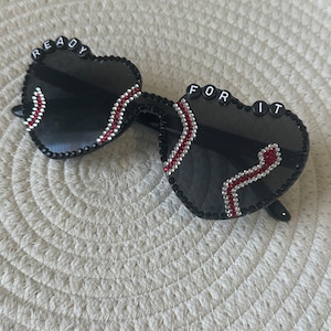 May include: Black heart-shaped sunglasses with rhinestones and red accents. The sunglasses have the words "READY FOR IT" spelled out on the frames.