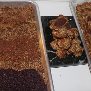 May include: Three baking sheets with different stages of granola bars. One sheet has a layer of dark brown granola, another has a layer of dark brown granola topped with a layer of dark purple seeds, and the third sheet has a pile of granola bars with a chocolate center.