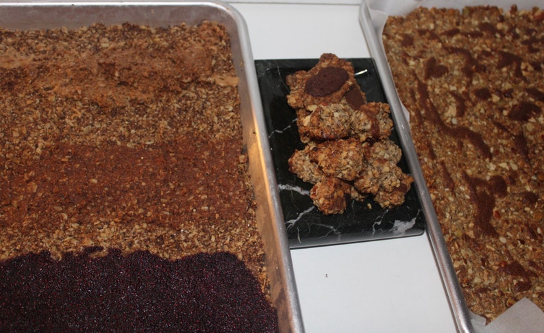 May include: Three baking sheets with different stages of granola bars. One sheet has a layer of dark brown granola, another has a layer of dark brown granola topped with a layer of dark purple seeds, and the third sheet has a pile of granola bars with a chocolate center.