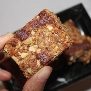 May include: A close-up of a granola bar with a brown, slightly glossy top. The bar is made with nuts, seeds, and dried fruit. It is held in a hand.