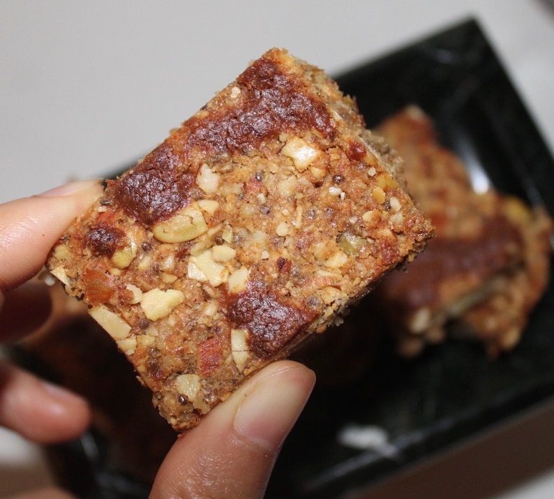 May include: A close-up of a granola bar with a brown, slightly glossy top. The bar is made with nuts, seeds, and dried fruit. It is held in a hand.