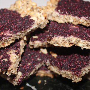 May include: A stack of homemade granola bars with a purple berry topping. The bars are rectangular and have a slightly crumbly texture.