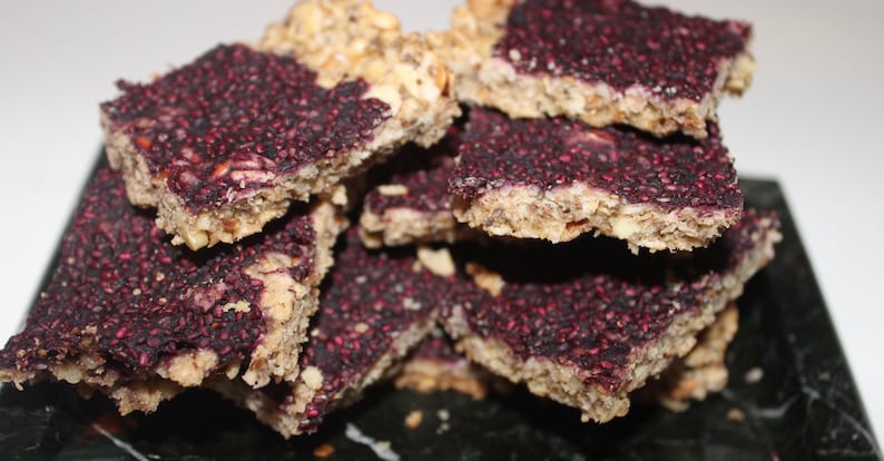 May include: A stack of homemade granola bars with a purple berry topping. The bars are rectangular and have a slightly crumbly texture.