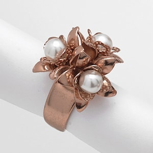 Puede incluir: Un anillo de oro rosa con un grupo de cuentas blancas con forma de perlas en el centro. El anillo está diseñado para parecer una flor con múltiples pétalos.