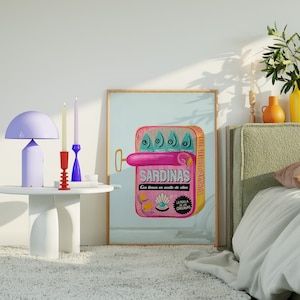 Op de afbeelding: Een vintage-stijl illustratie van een sardineblik met het woord "SARDINAS" erop gedrukt. Het blik is roze en heeft een blauw en wit ontwerp. De illustratie is ingelijst en hangt aan een witte muur.