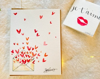 May include: A watercolor card with a white background and a yellow envelope filled with red and pink hearts. Hearts are also scattered around the envelope. The card is signed "Sophia". A separate card with a white background has the text "je t'aime" in black ink with a red lipstick kiss mark. The card is signed "Sophia".