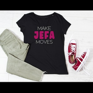 May include: A black V-neck t-shirt with the text MAKE JEFA MOVES in white and magenta, paired with olive green denim jeans and a pair of red canvas sneakers with white laces and white rubber soles, arranged on a light-colored wooden surface.