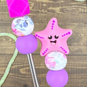 May include: Two decorative drink stirrers. One has a pink starfish with a smiling face, and the other has a pink square. Both have floral patterned and purple beads. The stirrers are on a wooden surface.