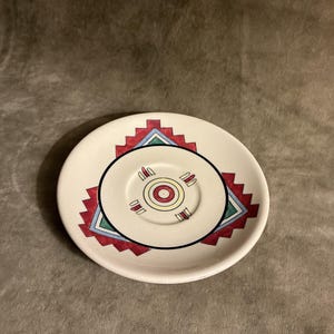 May include: A round ceramic saucer with a cream-colored base featuring a central red, yellow, and black circular motif. The design includes a geometric pattern with red jagged edges and teal triangles surrounding the center, creating a bold look.