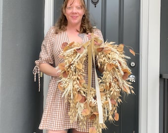 May include: A circular wreath composed of dried wheat stalks, beige grasses, and brown leaves, accented with small white berries. A long, smooth, light tan ribbon is tied in a bow at the top, with two long tails hanging down the center of the wreath.