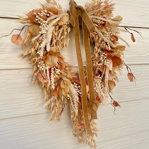 May include: A heart-shaped wreath constructed from dried grasses, beige wheat stalks, and rust-colored leaves. The arrangement features small white berries on thin stems and is accented with a long, hanging brown velvet ribbon tied at the top center.